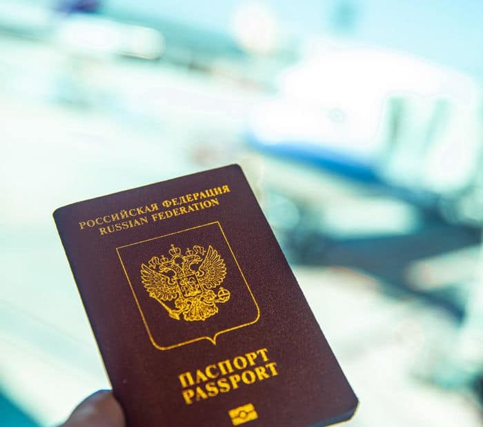 Close-up of a Russian passport being held near an airport gate window with blurred aircraft in the background.
